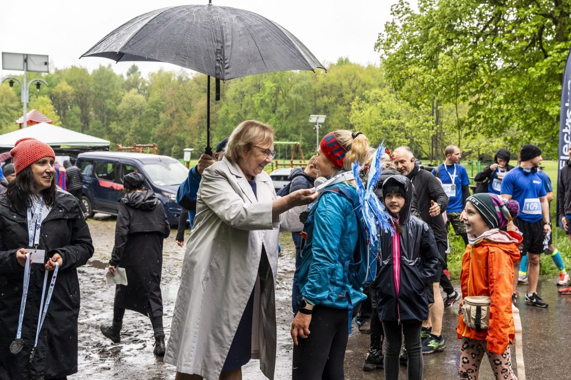 Zawodniczka odbiera medal na mecie, obok uczestnicy biegu w deszczu, jedna z osób trzyma parasol