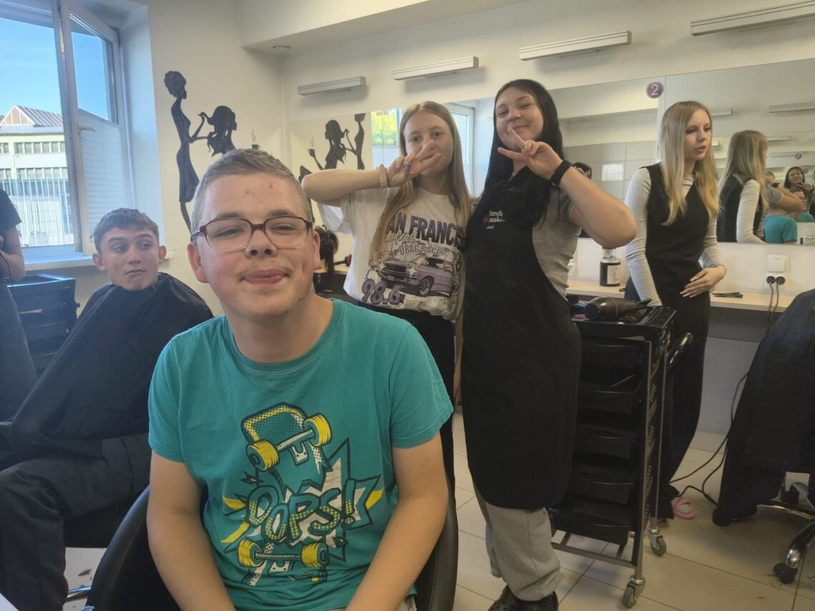 Uśmiechnięty uczeń w salonie fryzjerskim CKP, za nim uczennice klasy fryzjerskiej